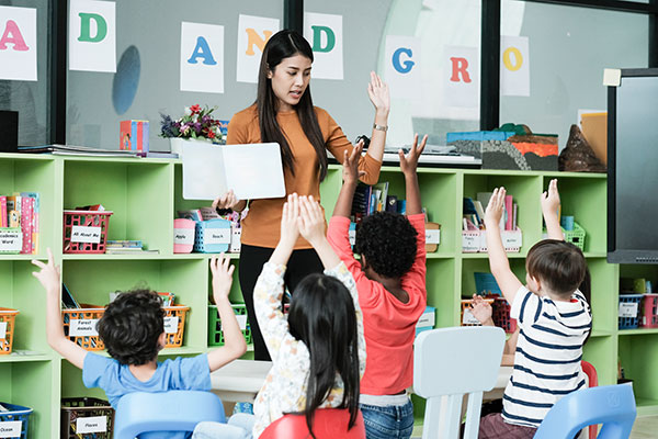 Teacher standing before a group of children's with their hands raised